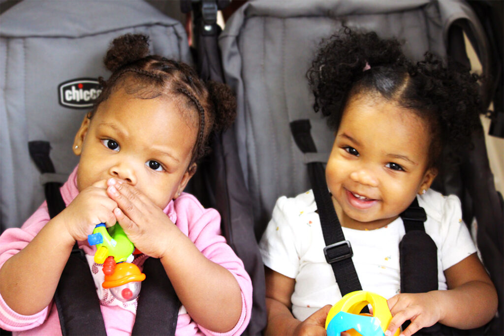 two little children in a stroller smiling at the camera, both holding toys.